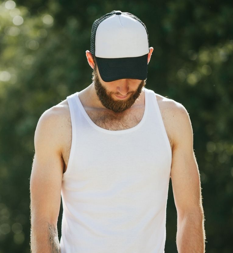 Young Caucasian man wearing a white tank top and a black-and-white cap, standing outdoors with arms relaxed